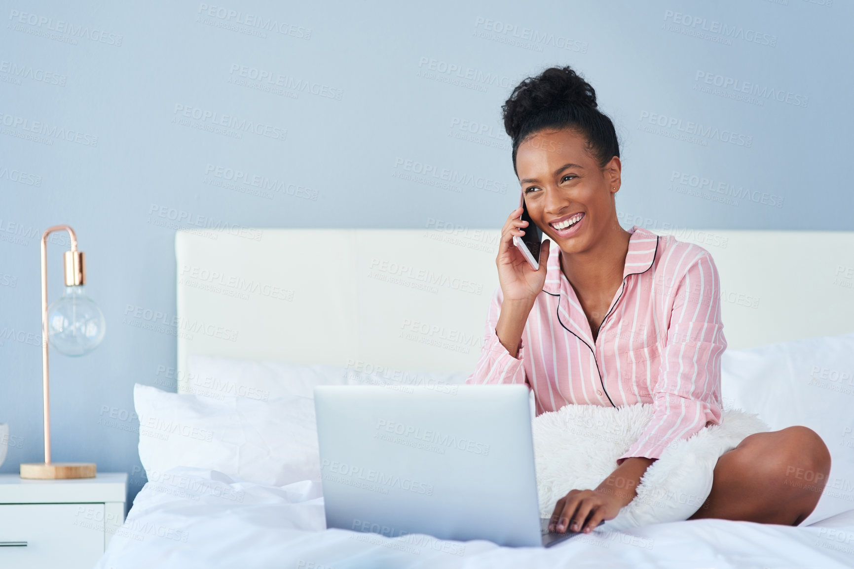 Buy stock photo Shot of an attractive young woman taking a phone call while using her laptop in bed at home