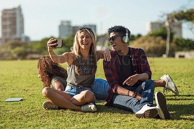 Buy stock photo Shot of two cheerful young friends taking a self portrait together while listening to music on headphones outside in a park