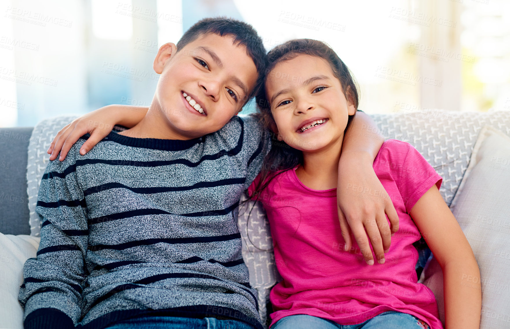 Buy stock photo Portrait of two adorable young siblings posing with their arms around each other while relaxing on a sofa at home