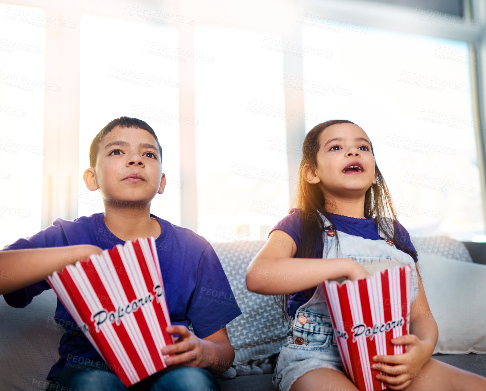 Buy stock photo Shot of two young children sitting on a sofa and eating popcorn while watching movies at home