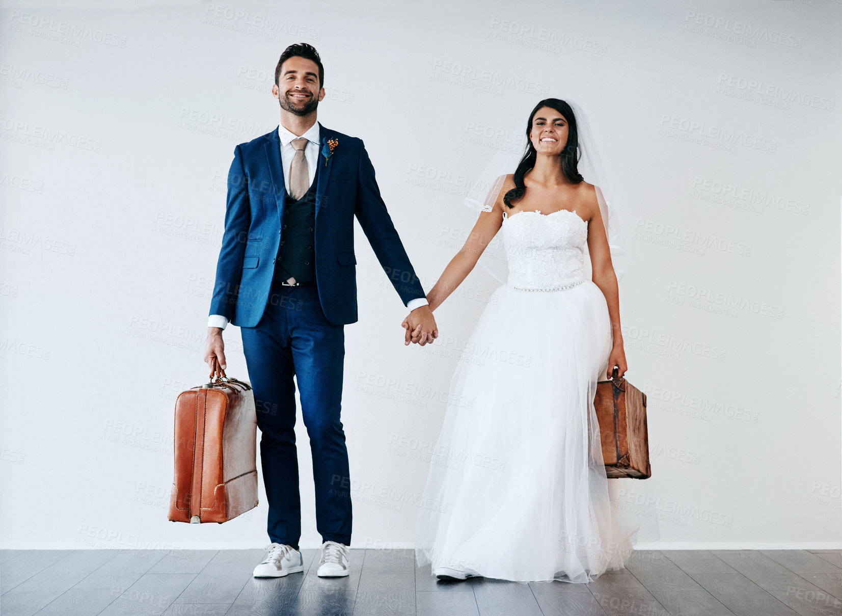 Buy stock photo Studio shot of a newly married couple holding hand and carrying bags while standing against a gray background