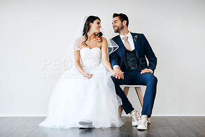 Buy stock photo Studio shot of a newly married young couple sitting together on a bench against a gray background