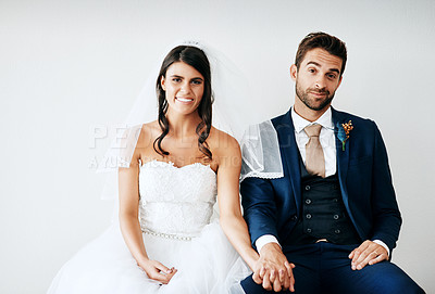 Buy stock photo Studio shot of a newly married young couple sitting together on a bench against a gray background