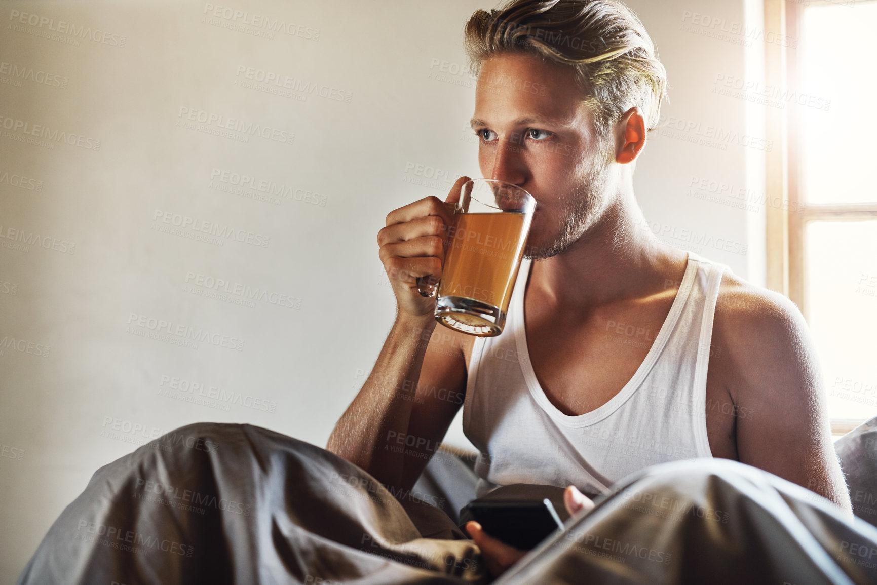 Buy stock photo Shot of a handsome young man using his cellphone while enjoying his morning tea in bed at home