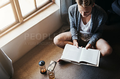 Buy stock photo High angle shot of a young man reading a book while sitting on the floor at home
