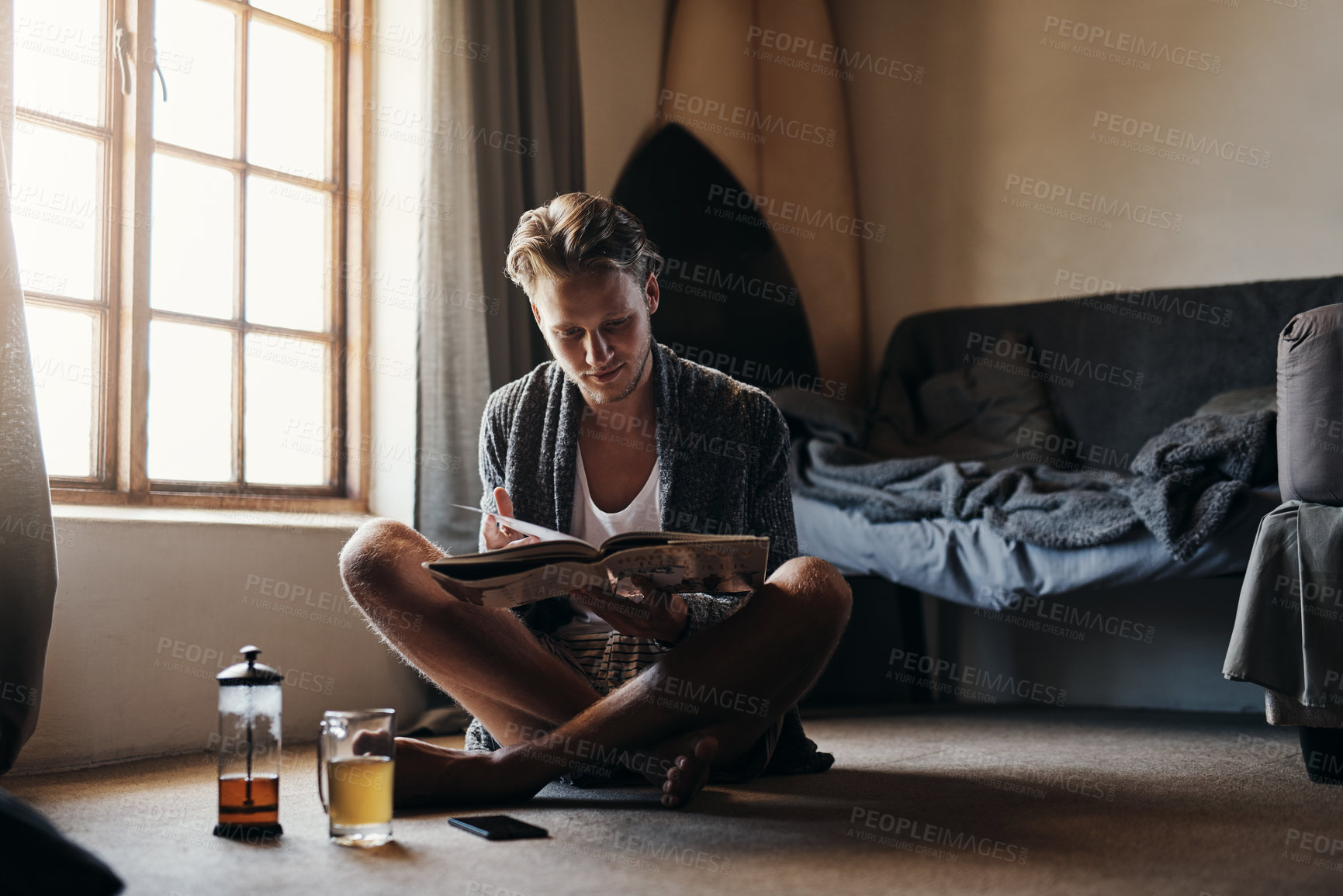 Buy stock photo Shot of a young man reading a book while sitting on the floor at home