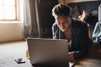 Buy stock photo Shot of a handsome young man using his laptop while lying down on the floor at home