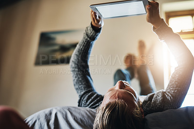 Buy stock photo Low angle shot of a young man using his digital tablet while relaxing in bed at home
