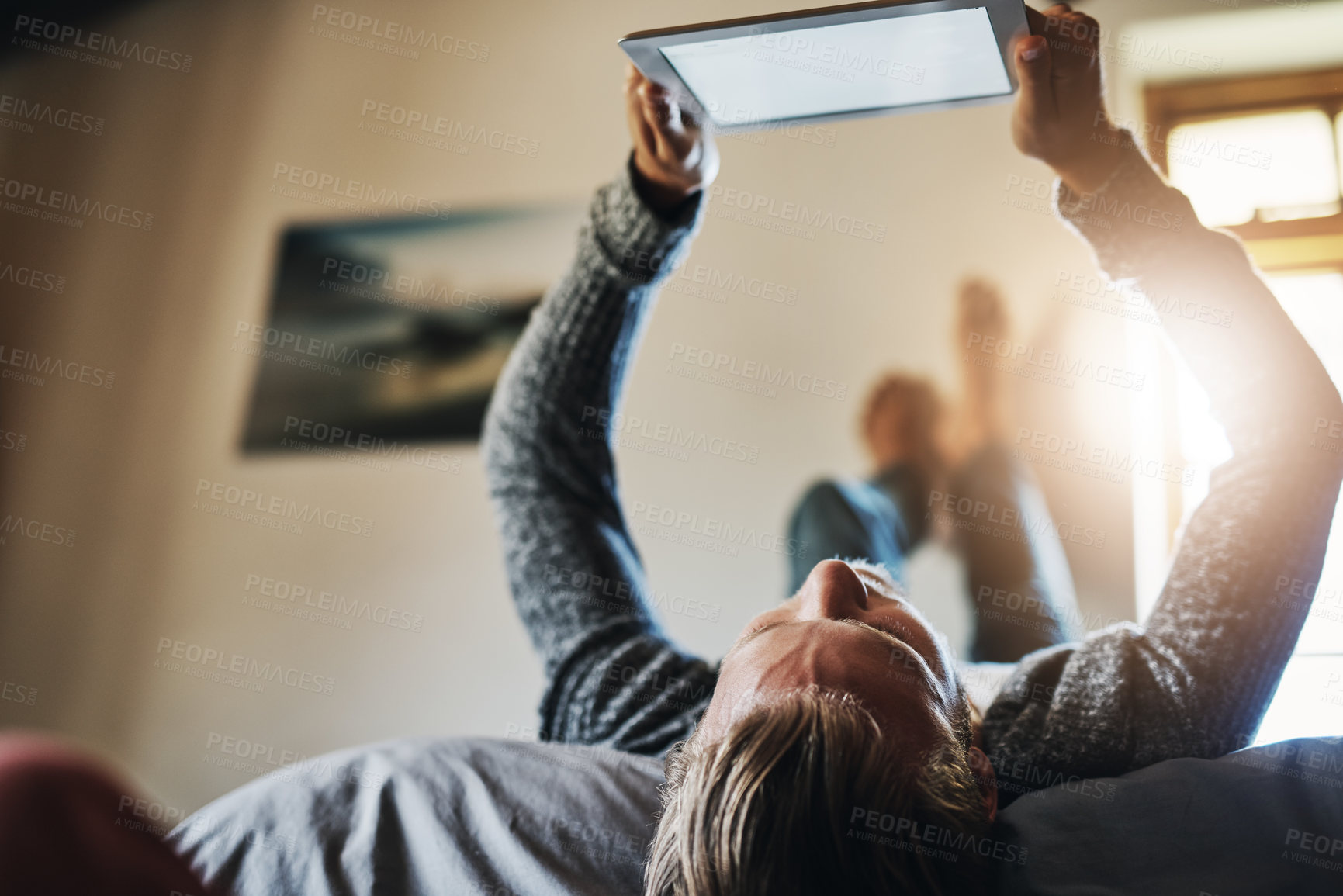 Buy stock photo Low angle shot of a young man using his digital tablet while relaxing in bed at home