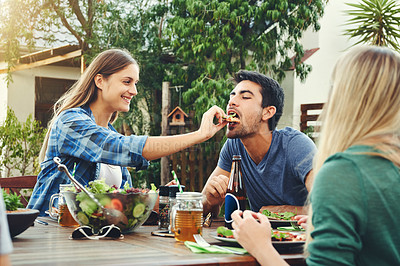 Buy stock photo Shot a cheerful young man being fed pizza by his girlfriend in an outdoor gathering amongst friends