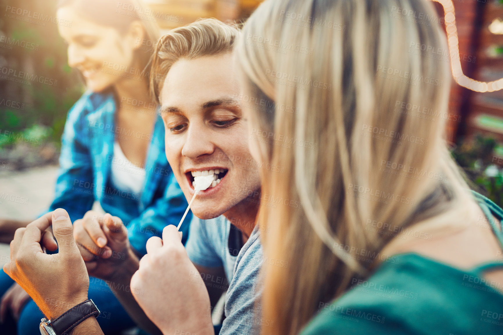 Buy stock photo Shot of a young cheerful man being fed a marshmallow by his girlfriend while relaxing outside with friends