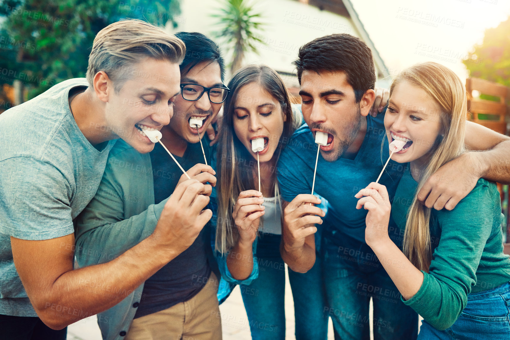 Buy stock photo Shot of a group of young friends posing together while eating marshmallows on sticks outside