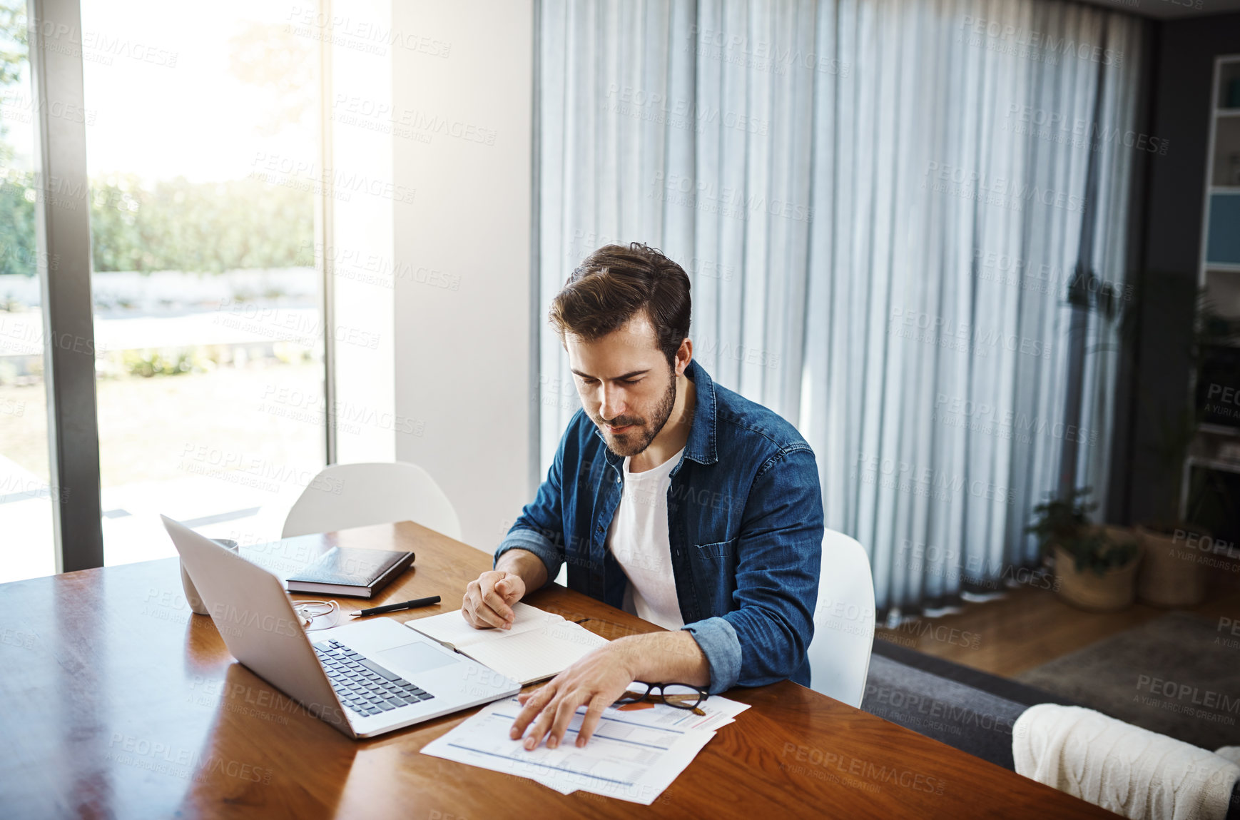 Buy stock photo Shot of a handsome young businessman sitting down and using his laptop while working from home