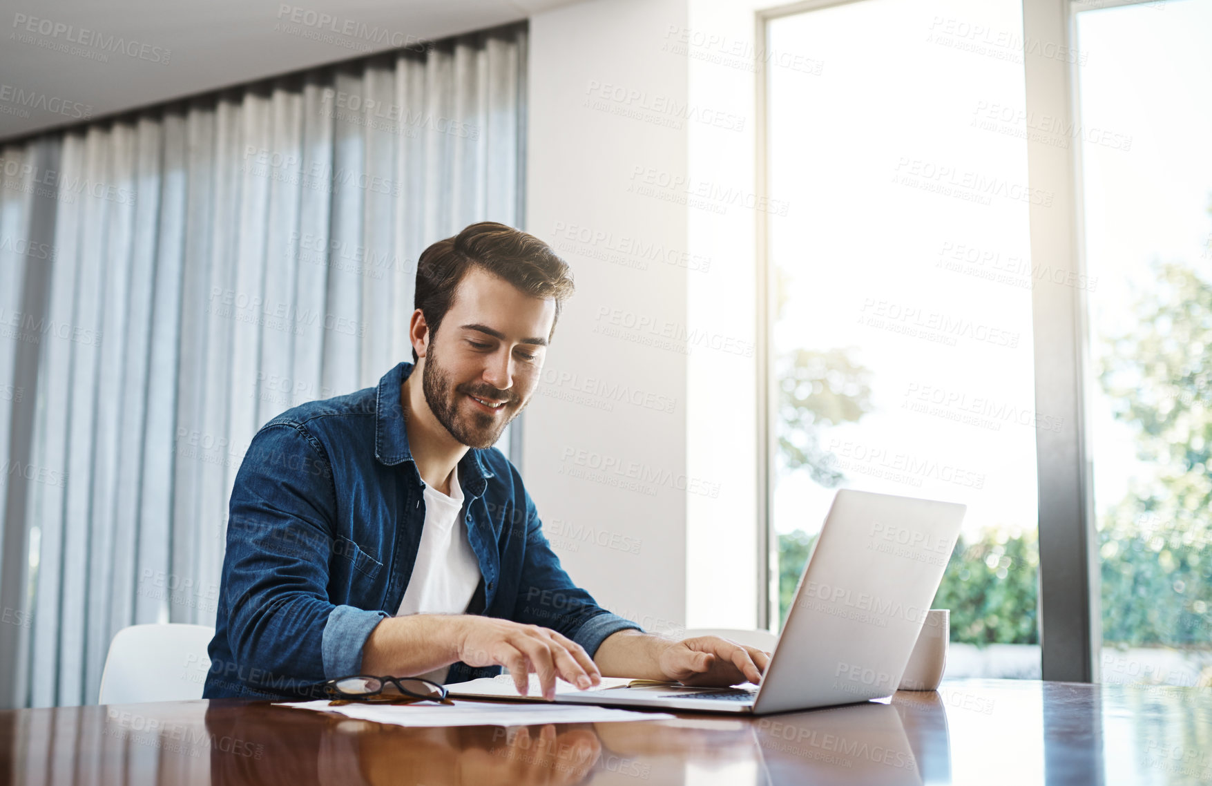 Buy stock photo Shot of a handsome young businessman sitting down and using his laptop while working from home