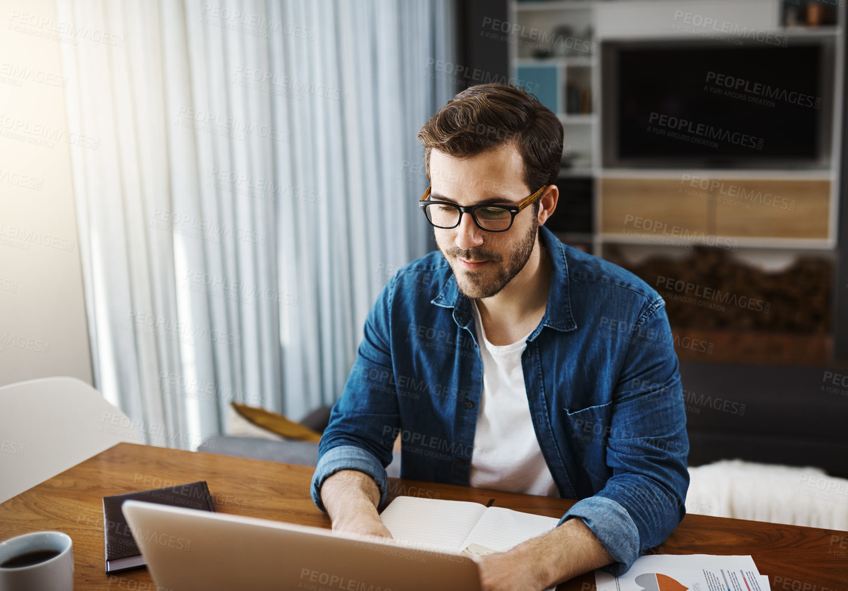 Buy stock photo Shot of a handsome young businessman sitting down and using his laptop while working from home