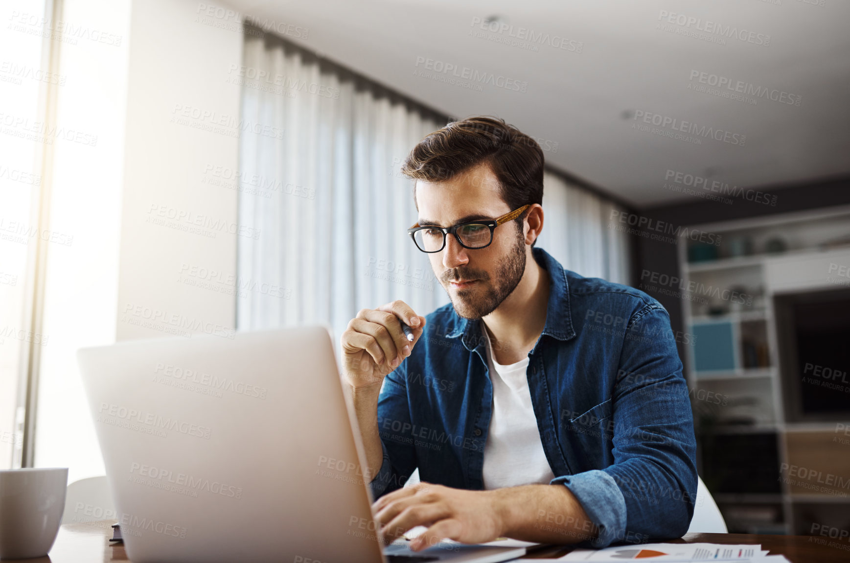 Buy stock photo Shot of a handsome young businessman sitting down and using his laptop while working from home