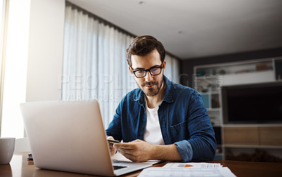 Buy stock photo Shot of a handsome young businessman sitting down and using his laptop to do some online shopping while working from home
