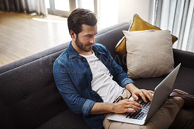 Buy stock photo Shot of a handsome young man using his laptop while relaxing on a sofa at home