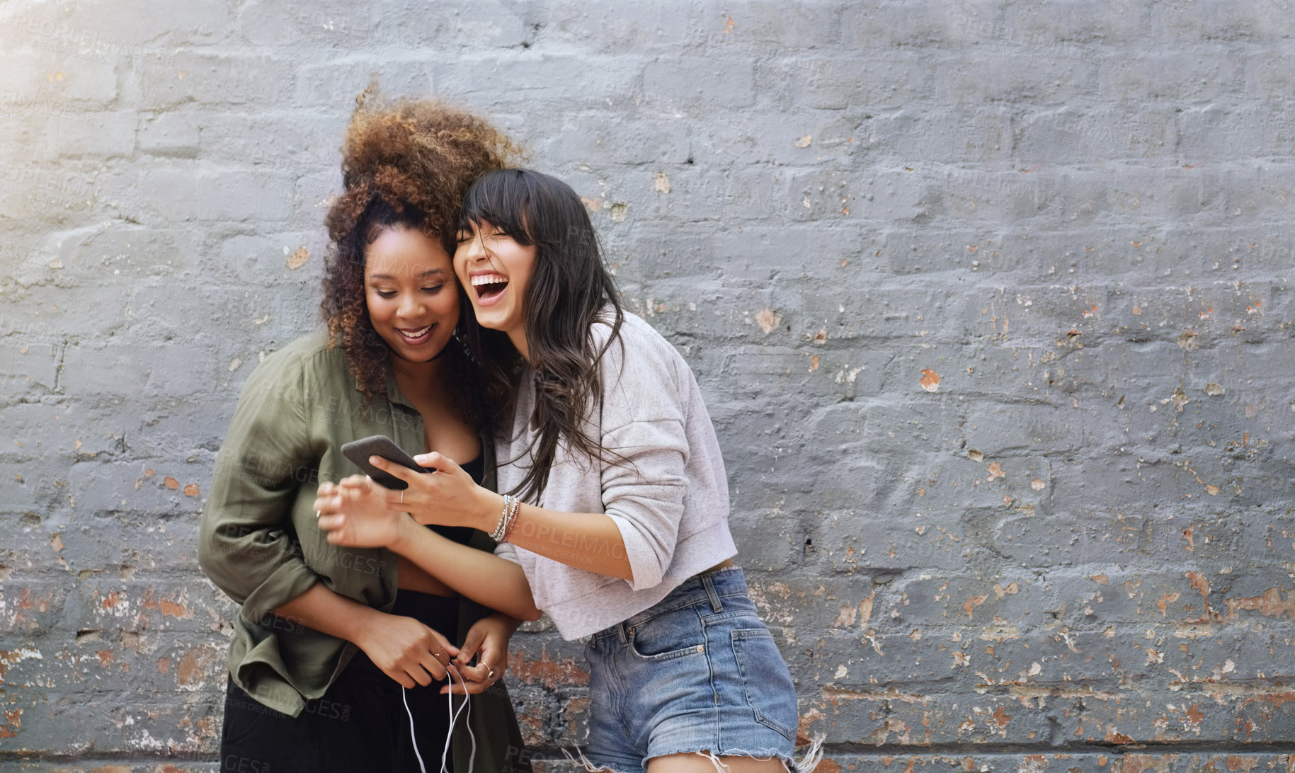 Buy stock photo Shot of two cheerful young women using a cellphone together while posing outdoors against a grey brick wall