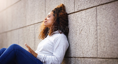 Buy stock photo Shot of an attractive young woman sitting down and leaning against a building while listening to music in the city