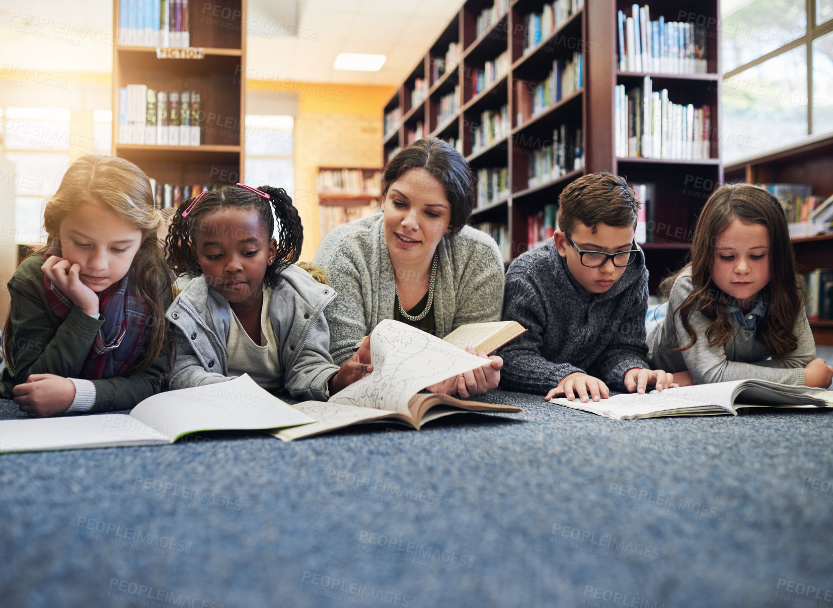Buy stock photo Shot of a group of elementary school kids and their teacher lying down while working in the library