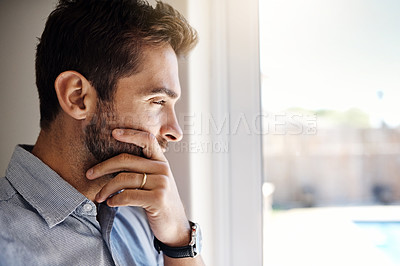 Buy stock photo Shot of a focused young man looking through a window while contemplating inside at home during the day
