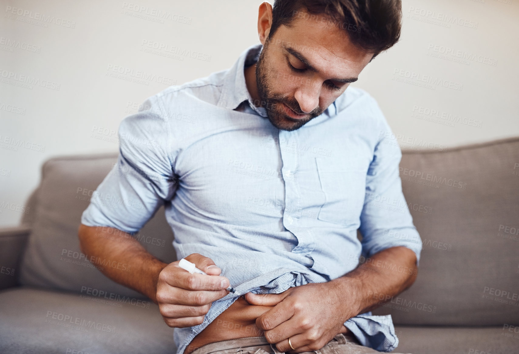 Buy stock photo Shot of a focused young man taking his daily insulin shot while being seated on a couch at home