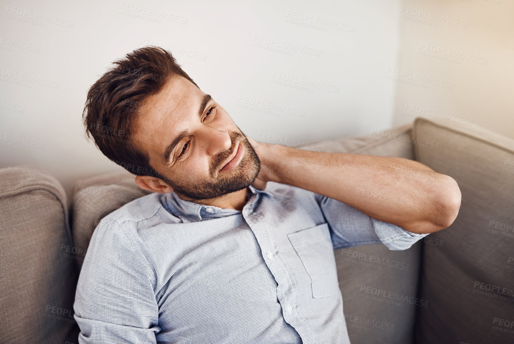 Buy stock photo Shot of a handsome young man holding his neck in pain while relaxing on a sofa at home