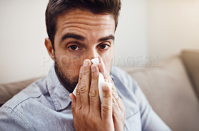 Buy stock photo Portrait of a handsome young man blowing his nose with a tissue while relaxing on a sofa at home