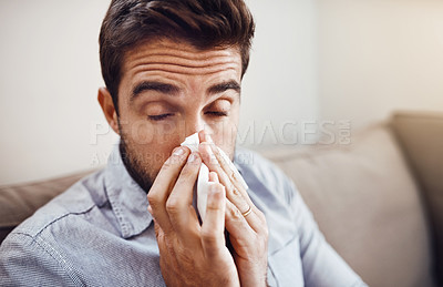 Buy stock photo Shot of a handsome young man blowing his nose with a tissue while relaxing on a sofa at home