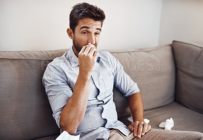 Buy stock photo Shot of a handsome young man blowing his nose with a tissue while relaxing on a sofa at home