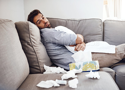Buy stock photo Portrait of a sickly young man holding a pillow while sitting on a couch with tissues laying all around at home