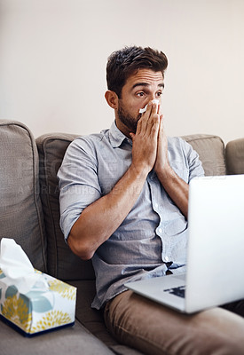 Buy stock photo Shot of a young businessman blowing his nose with a tissue while trying to work on his laptop at home
