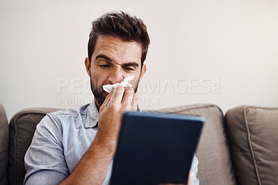 Buy stock photo Shot of a sickly young man blowing his nose with a tissue while browsing through his digital tablet at home