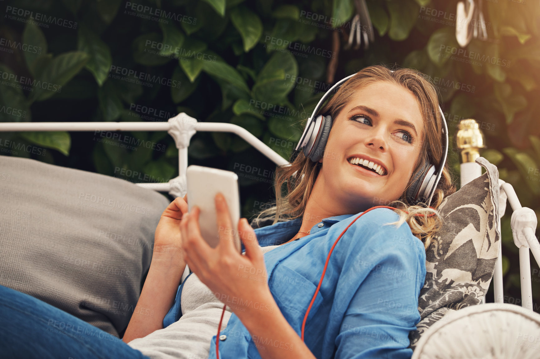 Buy stock photo Shot of a young woman using headphones with her smartphone while relaxing in the garden at home