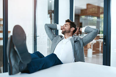 Buy stock photo Shot of a handsome young businessman relaxing with his feet up on a desk in a modern office