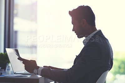 Buy stock photo Cropped shot of a handsome young businessman using a laptop and cellphone at his desk in a modern office