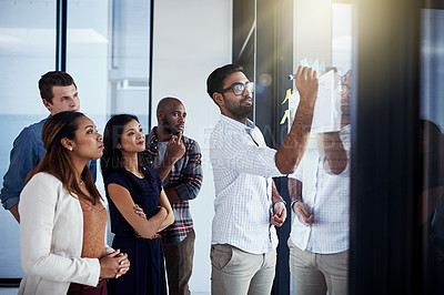 Buy stock photo Shot of a young businessman giving a demonstration on a glass wall to his colleagues in a modern office
