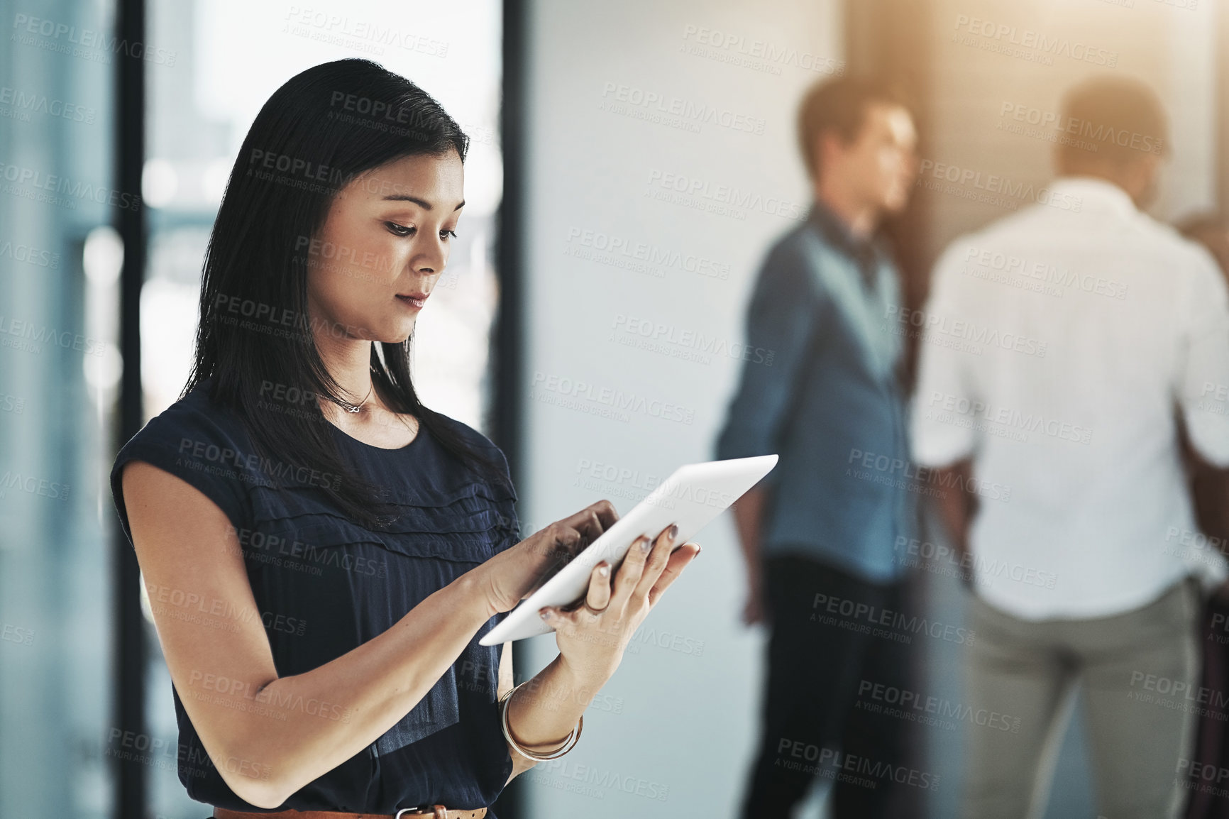 Buy stock photo Shot of a young businesswoman using her digital tablet in a modern office with her colleagues in the background