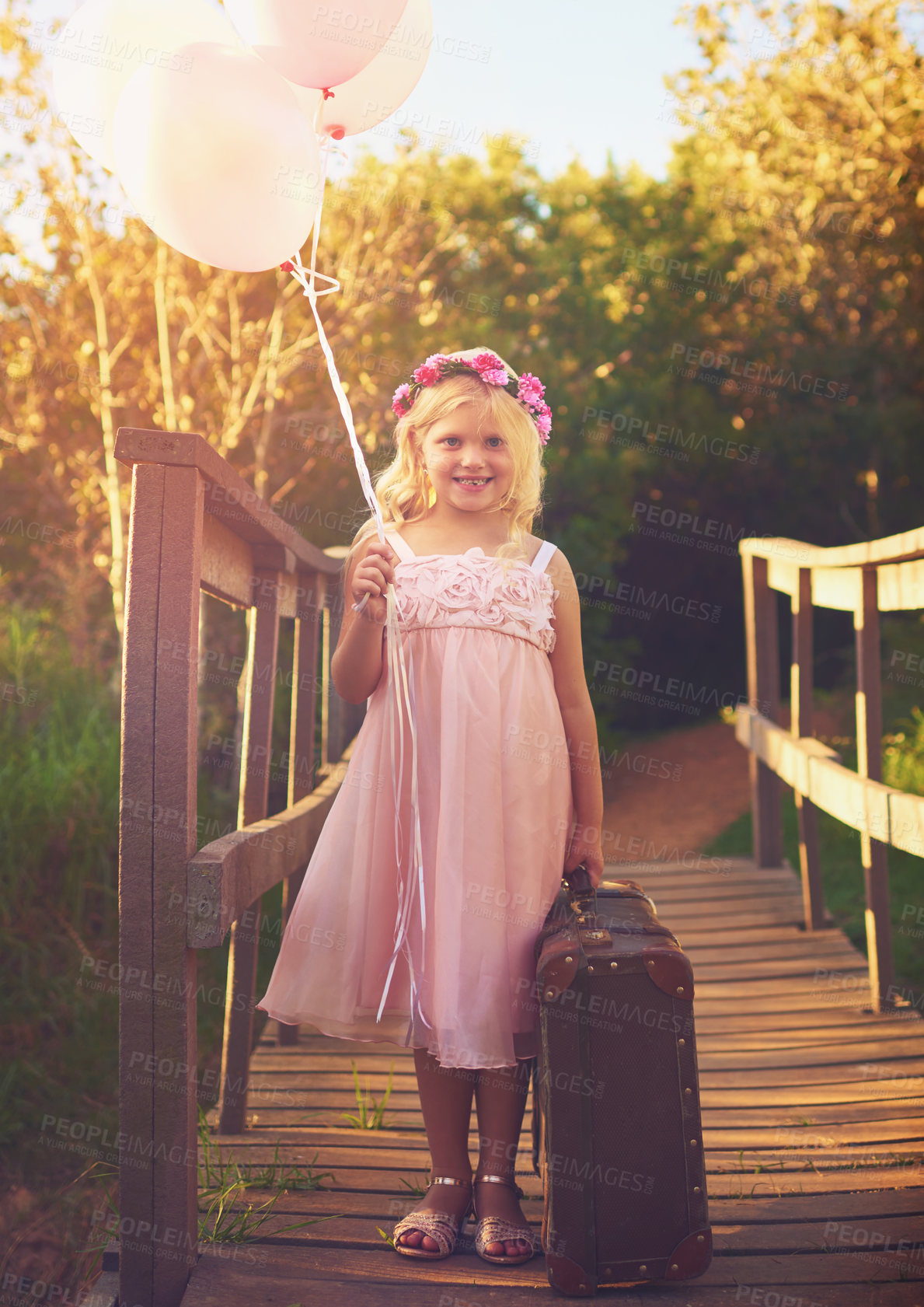 Buy stock photo Shot of a happy little girl holding balloons and a suitcase while standing in the middle of a bridge