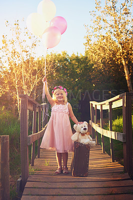Buy stock photo Shot of a happy little girl holding balloons and a teddy bear while standing in the middle of a bridge
