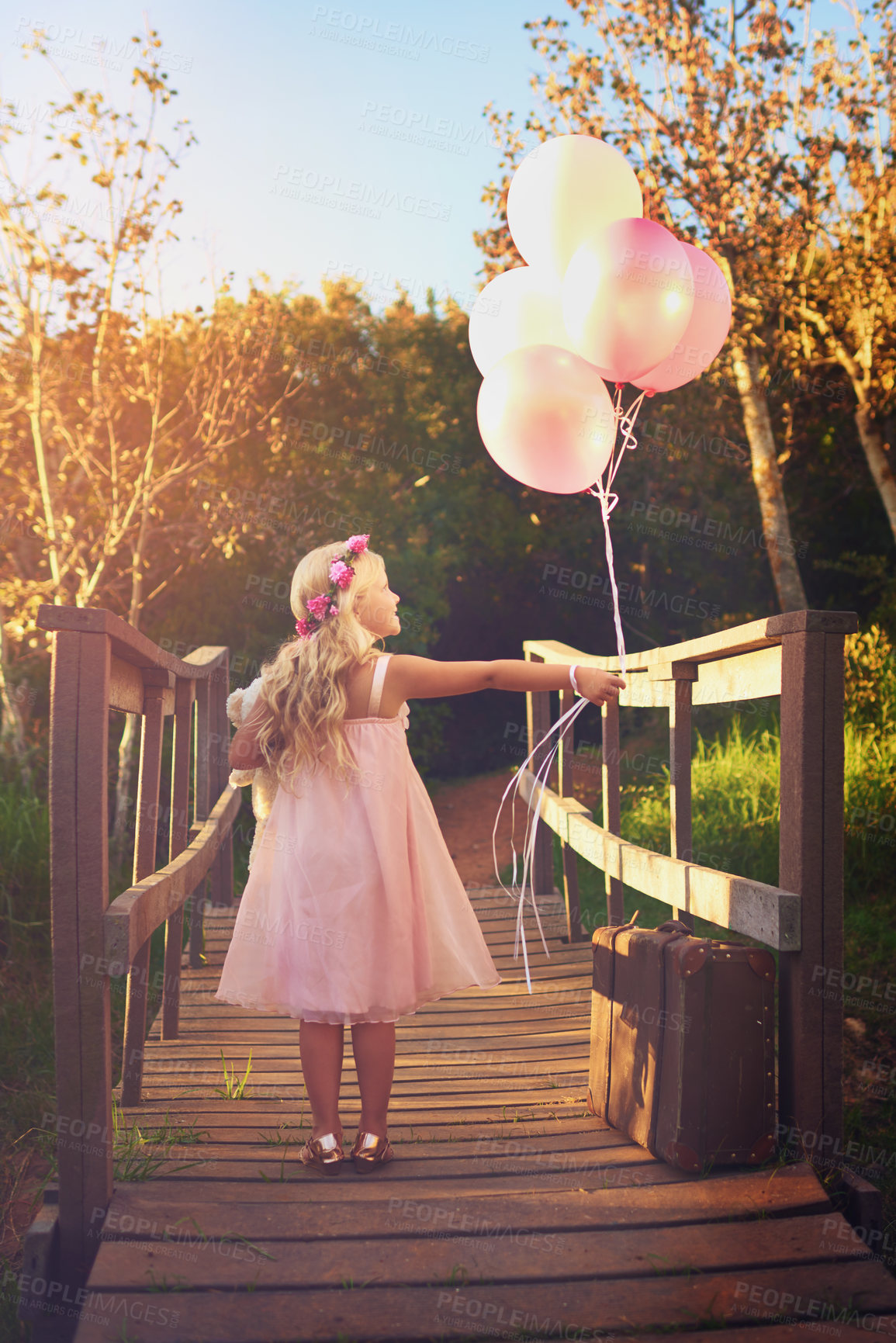 Buy stock photo Shot of a happy little girl holding balloons and a teddy bear while standing in the middle of a bridge
