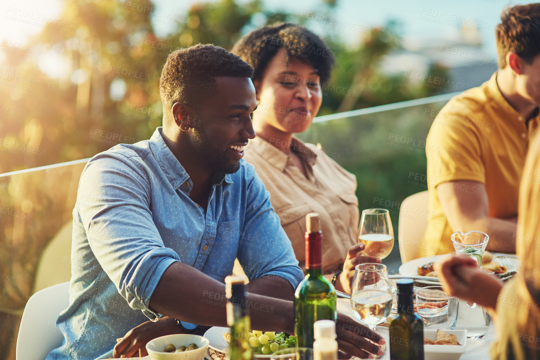 Buy stock photo Shot of cheerful young man  sitting around a table with friends at a gathering outdoors