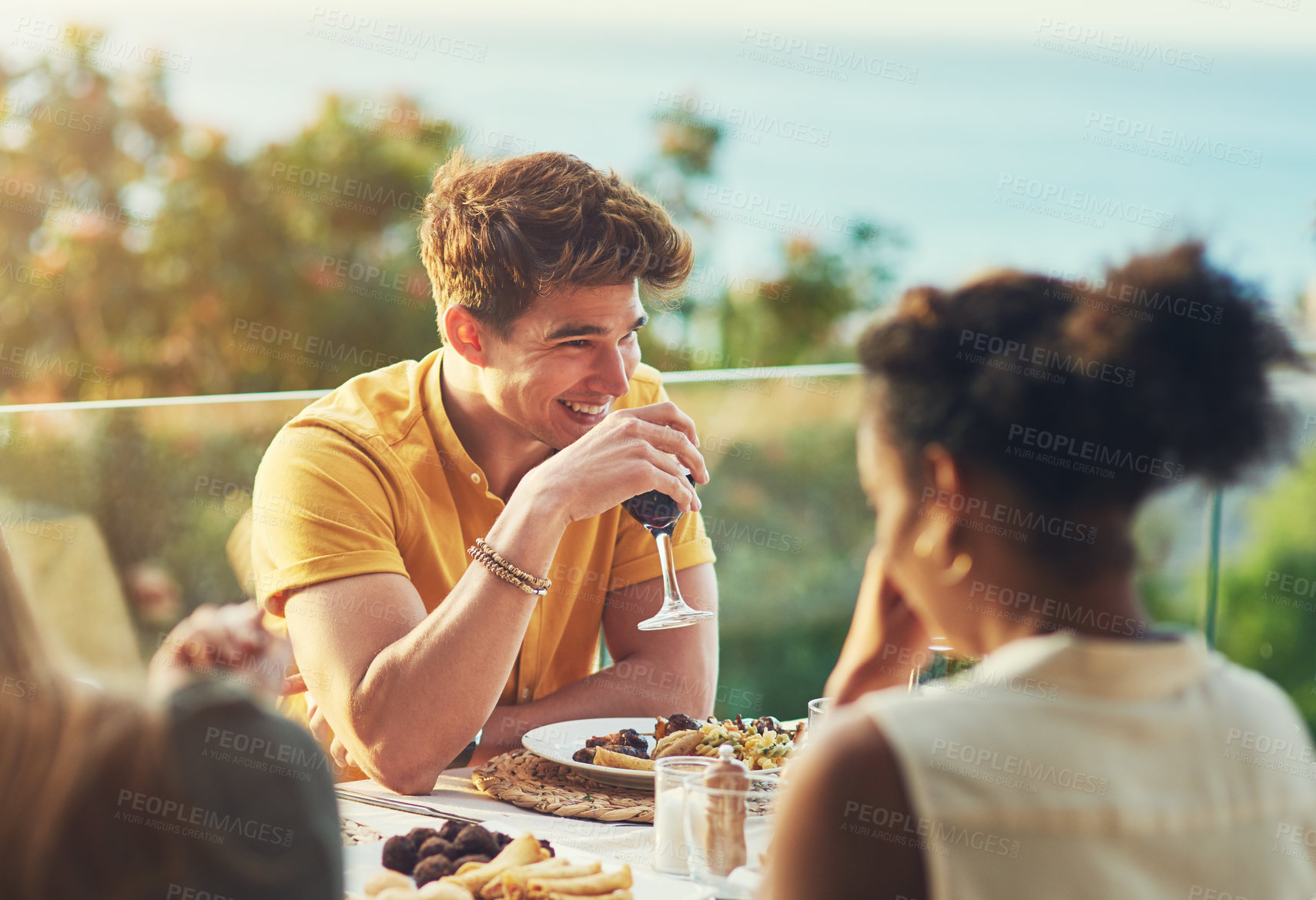 Buy stock photo Shot of a handsome young man enjoying a glass of wine while sitting around a table with friends outdoors