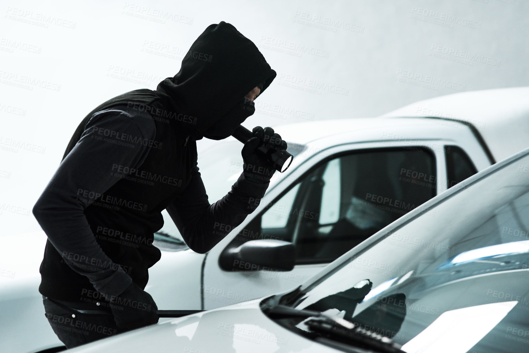 Buy stock photo Shot of a masked criminal using a torch light to look inside an empty car inside a parking lot