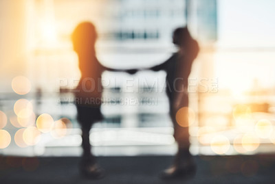 Buy stock photo Silhouetted shot of two unrecognizable businesswomen shaking hands while standing in a modern workplace