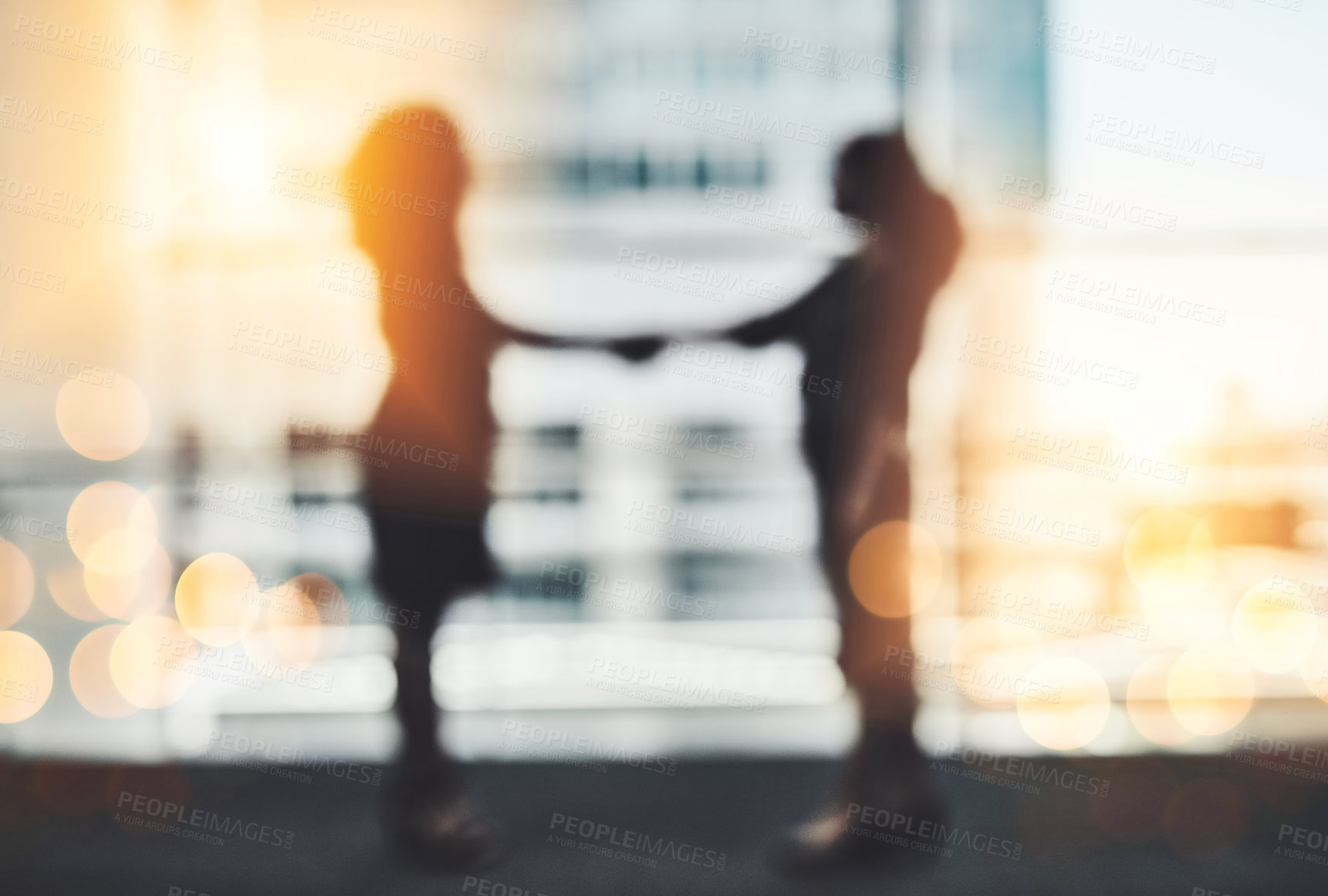 Buy stock photo Silhouetted shot of two unrecognizable businesswomen shaking hands while standing in a modern workplace