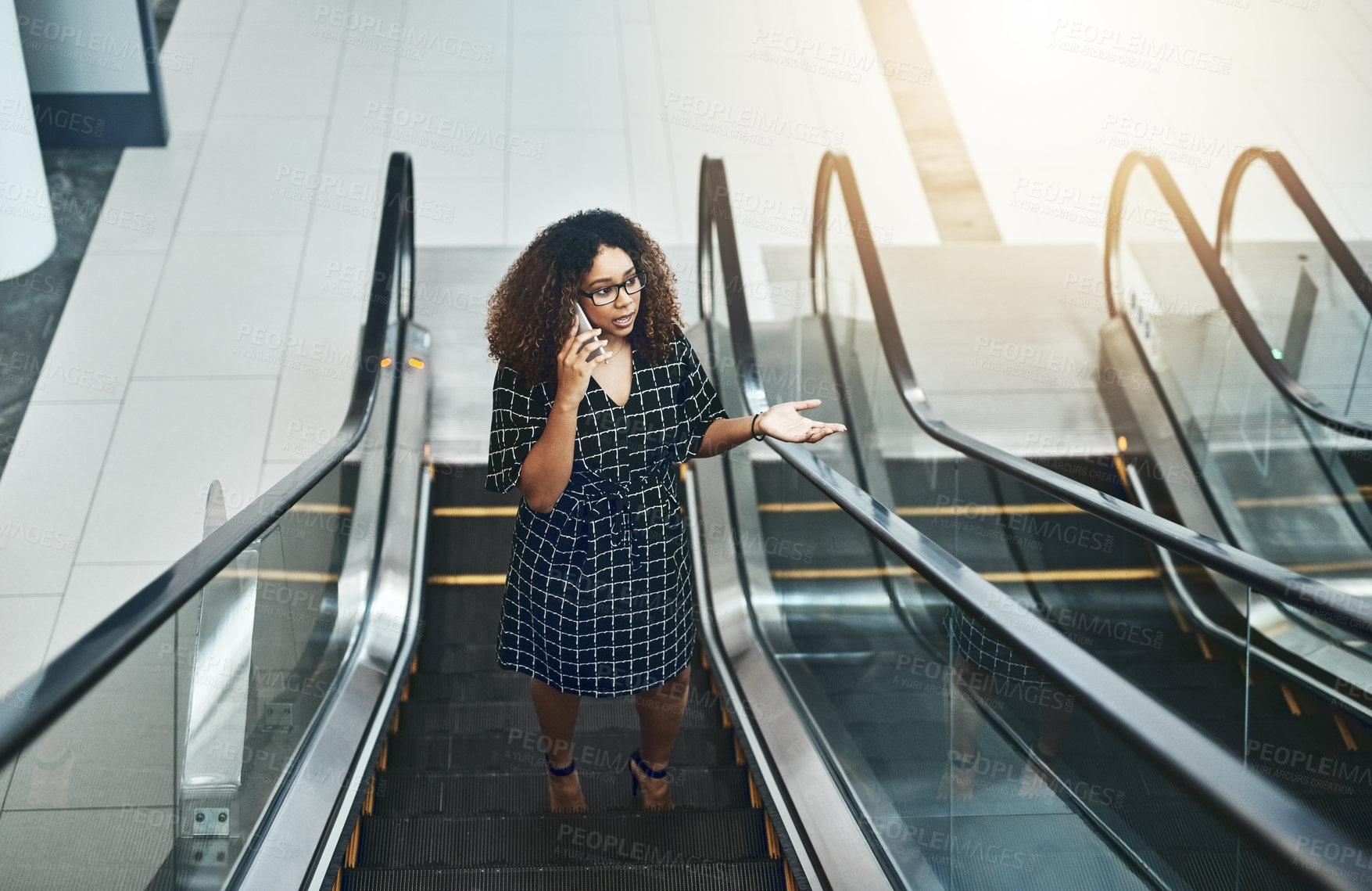 Buy stock photo High angle shot of an attractive young businesswoman taking a phonecall while going up an escalator in a modern office