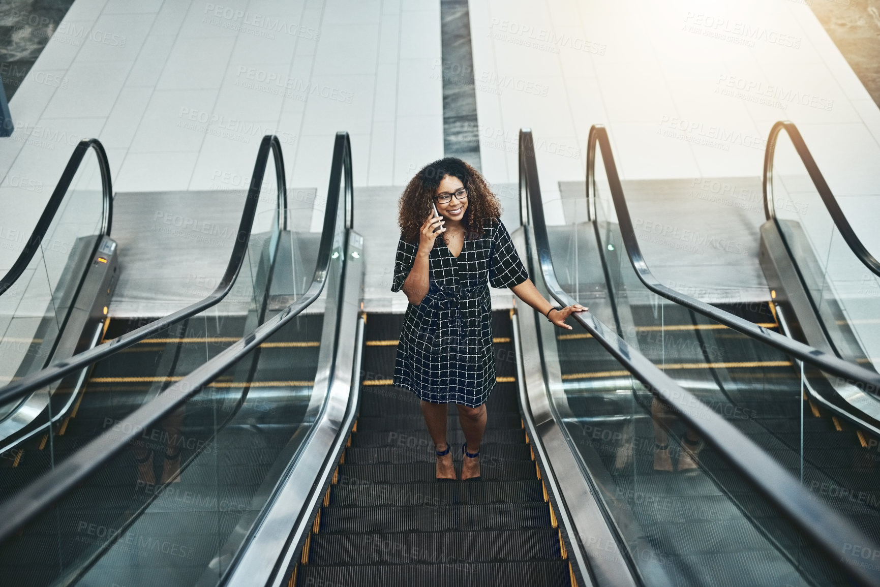 Buy stock photo High angle shot of an attractive young businesswoman taking a phonecall while going up an escalator in a modern office