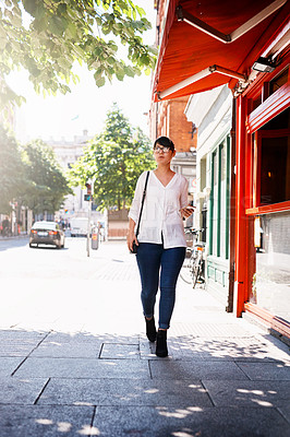 Buy stock photo Shot of a beautiful young woman walking on a sidewalk past shops outside in a town during the day